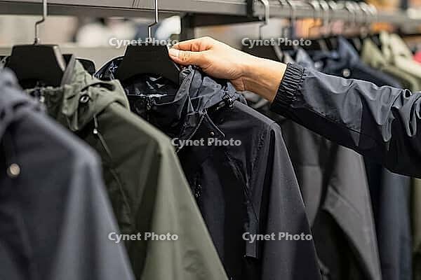 Technical outdoor apparel shopping defines functional garment curation, a hand inspects waterproof hooded jackets on rails inside an urban store, synthetic weather resistant fabrics define utilitarian trends in consumer culture, dark windbreakers line up  [IBR125502131]