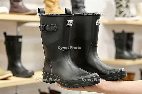 Waterproof black rubber boots held in hand at retail store, durable footwear display for outdoor protection and rainy weather equipment, functional wellington gumboots crafted from molded vulcanized polymer with side cinch buckle and lugged traction tread [IBR125502153]