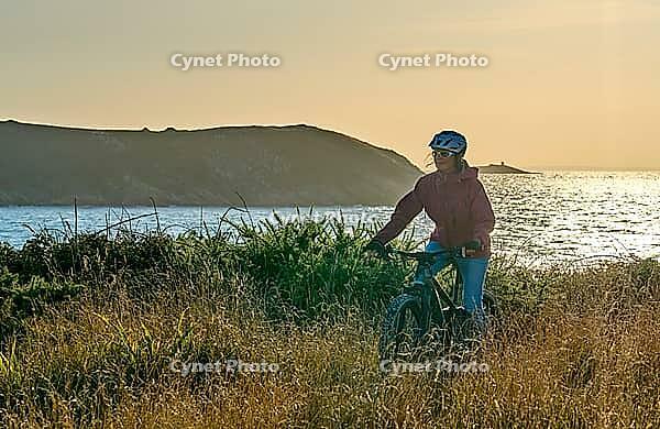 Active senior woman cycling with her electric mountain bike along the wild atlantic coast of Finisterre at Cape Frehel, Brittany, France [IBR125521080]