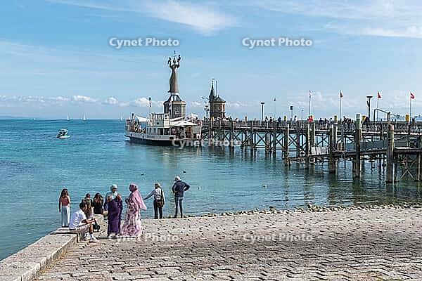 Visitors stand on the stone promenade and watch the ship at the sunny pier, harbor with the Imperia statue, Constance on Lake Constance, Baden-Württemberg, Germany [IBR125521084]