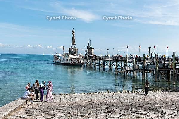 A small group gathers on the shore opposite an excursion boat and a pier reaching far into the sea, harbor with the Imperia statue, Constance on Lake Constance, Baden-Württemberg, Germany [IBR125521091]