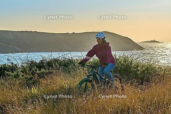 Active senior woman cycling with her electric mountain bike along the wild coast of Finisterre in southern Brittany next to Camaret-sur-Mer, France [IBR125521116]