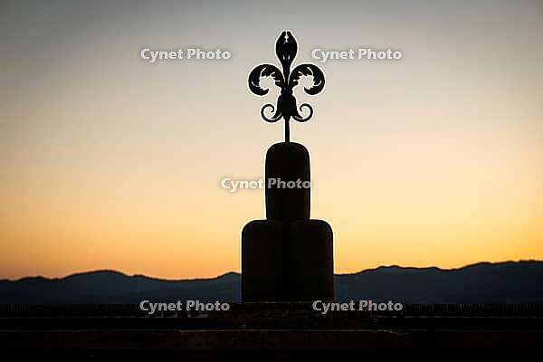 A Fleur-de-lis symbol, stylized lily, stands out as a dark silhouette against the orange sunset and a mountain range, Gubbio, Umbria, Italy [IBR125521160]