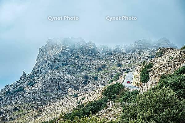 Rocky limestone mountain landscape with a winding paved road and small red car under misty, overcast sky in El Torcal near Antequera, Andalusia, Spain. Green shrubs and trees cover the slopes around the natural park roadway [IBR125527076]