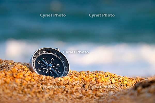 Conceptual photo, close-up of a compass lying in wet sand consisting of small crumbs of shells, against the backdrop of a blue calm sea [IBR125527077]