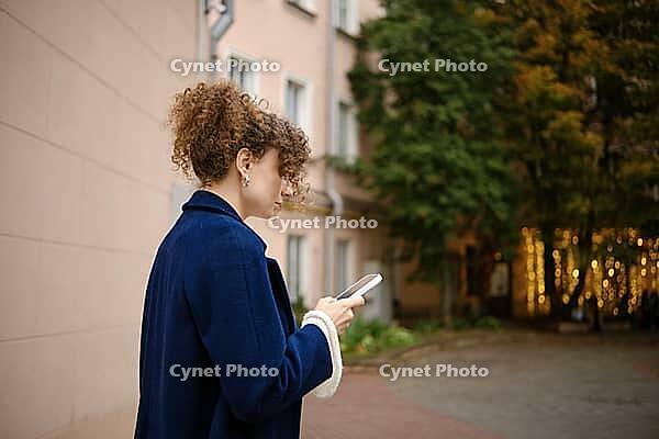 Woman using smartphone to order taxi on urban sidewalk beside apartment buildings and trees. Mobile communication, messaging and digital connectivity. Everyday city life and technology use [IBR125536432]