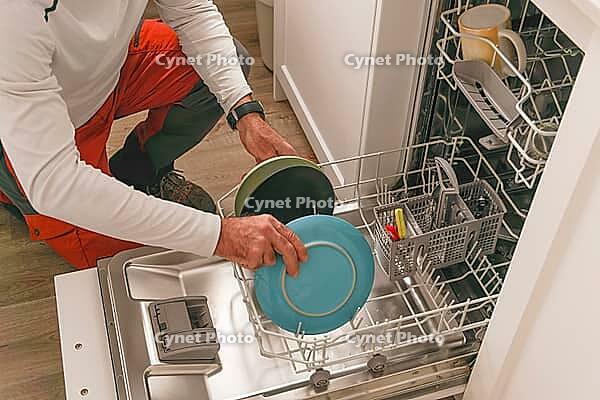 Man in a white t-shirt, the dishes in the dishwasher, in the kitchen of his house [IBR125536454]