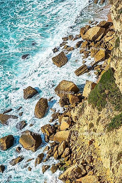 Rocky sea cliff coastline with large boulders, turquoise Atlantic water and breaking waves near Barbate in Andalusia, Spain. View from above shows textured sedimentary rock formations along the rugged shore in bright daylight [IBR125536461]