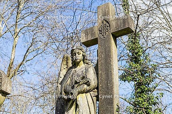 A weathered stone angel statue stands next to a large cross monument in the historic Highgate Cemetery in London under a blue sky [IBR125557550]