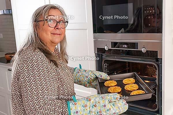 Elderly white-haired woman putting a tray of cookies into the ovens [IBR125557576]