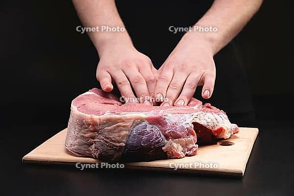 A man massages a fresh piece of beef with his hands on a wooden cutting board. Close up on black background [IBR125557615]