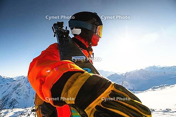 A guy in a ski suit holds skis in his hand and stands on a snowy mountain looking into the distance at the sun and a mountain range, ski resort and recreation development [IBR125565997]