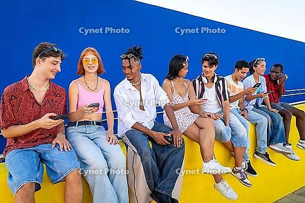 Diverse young adults sitting on a yellow wall, smiling and interacting with their smartphones against a vibrant blue background, symbolizing digital communication and social connection [IBR125566036]