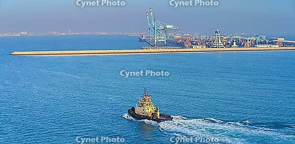 Manama, Bahrain - Jan 08 2024, Panoramic view of passing tugboat, a boat, in the background a commercial international port with cranes, pier, buildings and containers, in the daytime, Manama, Bahrain [IBR125566055]