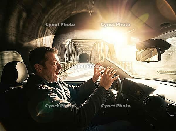 A man's startled face and raised hands fill the foreground, while a steel truss bridge and intense sunlight appear beyond the windshield, AI generated [IBR125592659]
