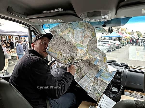 Inside the vehicle, a man grips a large folded road map across the dashboard as shoppers, market tents and parked cars line the street outside, AI generated [IBR125592684]