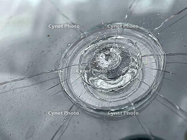 A close view of shattered automotive glass shows a central impact crater, concentric rings, fine spiderweb fractures, and scattered moisture, AI generated [IBR125592693]
