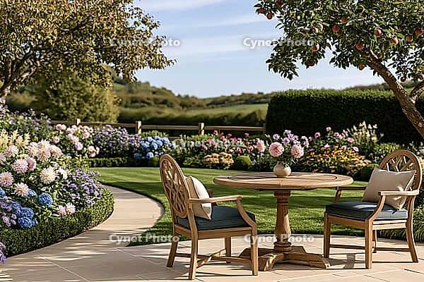 A sunlit garden patio with a wooden table and two chairs, surrounded by flowering borders, clipped hedges, and an apple tree. AI generated [IBR125592704]