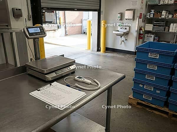 A stainless worktable holds a scale, probe thermometer, and receiving checklist beside reusable crates at a kitchen loading bay for food deliveries, AI generated [IBR125592715]