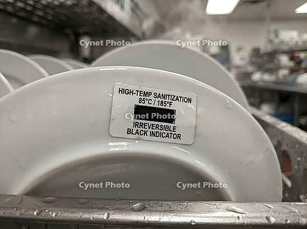 A water-speckled plate displays an irreversible heat indicator at 85degC, with dishware and stainless equipment visible in a commercial kitchen, AI generated [IBR125592724]