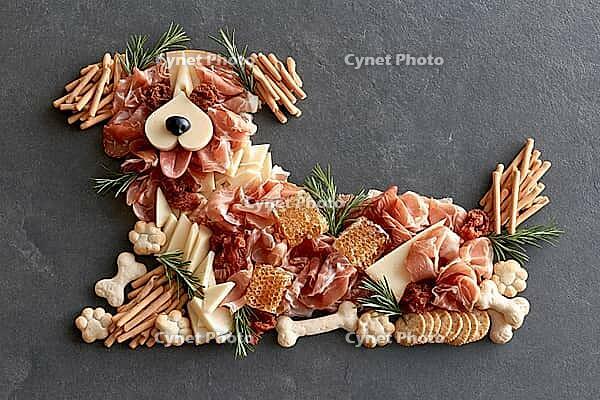 A playful dog-shaped charcuterie arrangement made from cured meats, cheese, breadsticks, crackers, nuts, and rosemary on a dark slate surface. AI generated [IBR125592737]