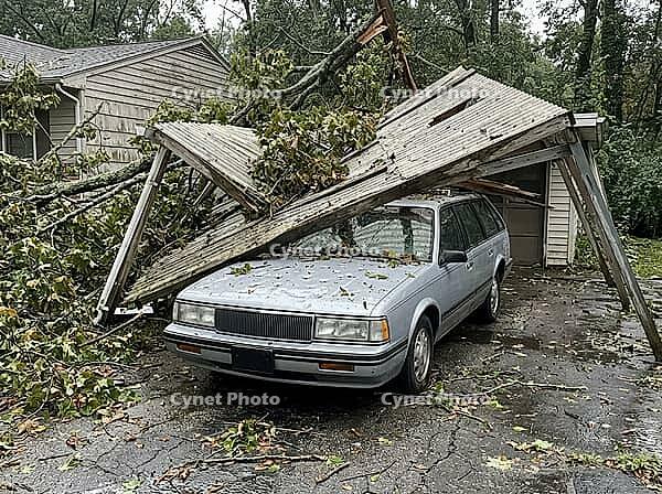 Broken branches and roofing panels cover the driveway around the vehicle, while a house and dense trees frame the residential scene, AI generated [IBR125592746]