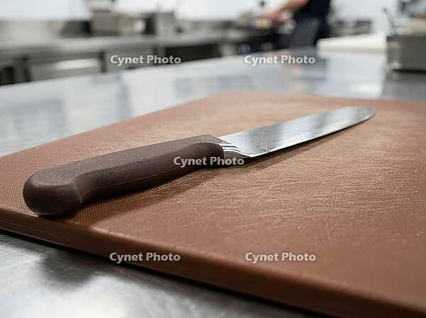 Close focus shows a brown prep board and polished blade on a stainless worktop, with kitchen equipment and a blurred worker nearby, AI generated [IBR125592748]