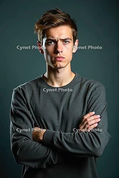 Portrait of a serious young man crossing his arms, looking upset and skeptical on a dark studio background, AI generated [IBR125621348]