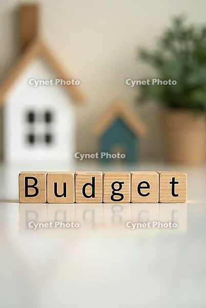 Wooden blocks spelling budget are reflecting on a white surface with miniature houses and a plant in the background, representing home budget planning, AI generated [IBR125621365]
