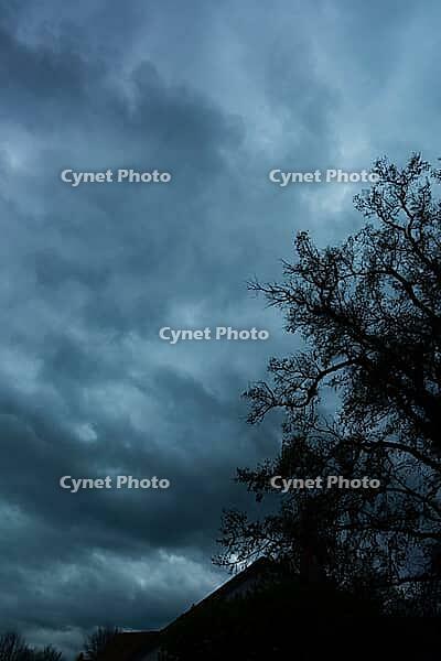 A bare tree and rooftop are silhouetted beneath heavy, dark blue-gray clouds [IBR125657365]