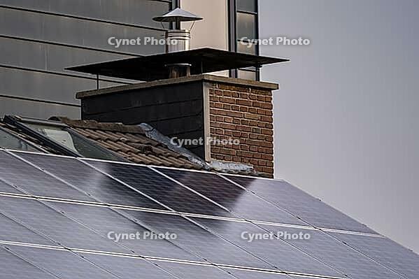 Solar panels cover a house roof beside a brick chimney and metal flue in daylight, with building walls visible in the background [IBR125657366]