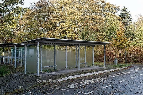 Metal shelters with glass panels stand beside a paved parking area, with fallen leaves on the ground and trees showing autumn colors in the background [IBR125657373]