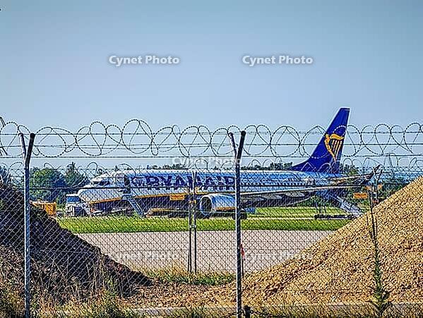 Airplane of the airline Ryanair, a white passenger aircraft with blue-yellow lettering and blue tail unit with yellow harp standing on the apron and being handling with vehicles and gangways, plus a wire mesh fence with barbed wire in the foreground, pile [IBR125657384]