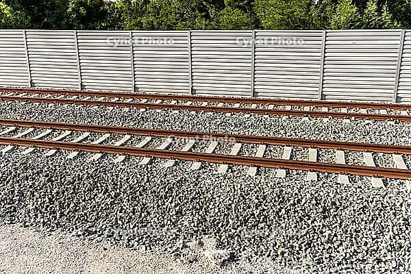 Railway tracks cross a gravel bed in front of a long sound barrier, with green treetops visible beyond [IBR125657403]
