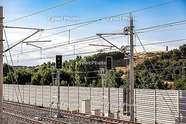 Railway signals and overhead electrical lines stand above tracks bordered by a sound wall and green hillside [IBR125657440]